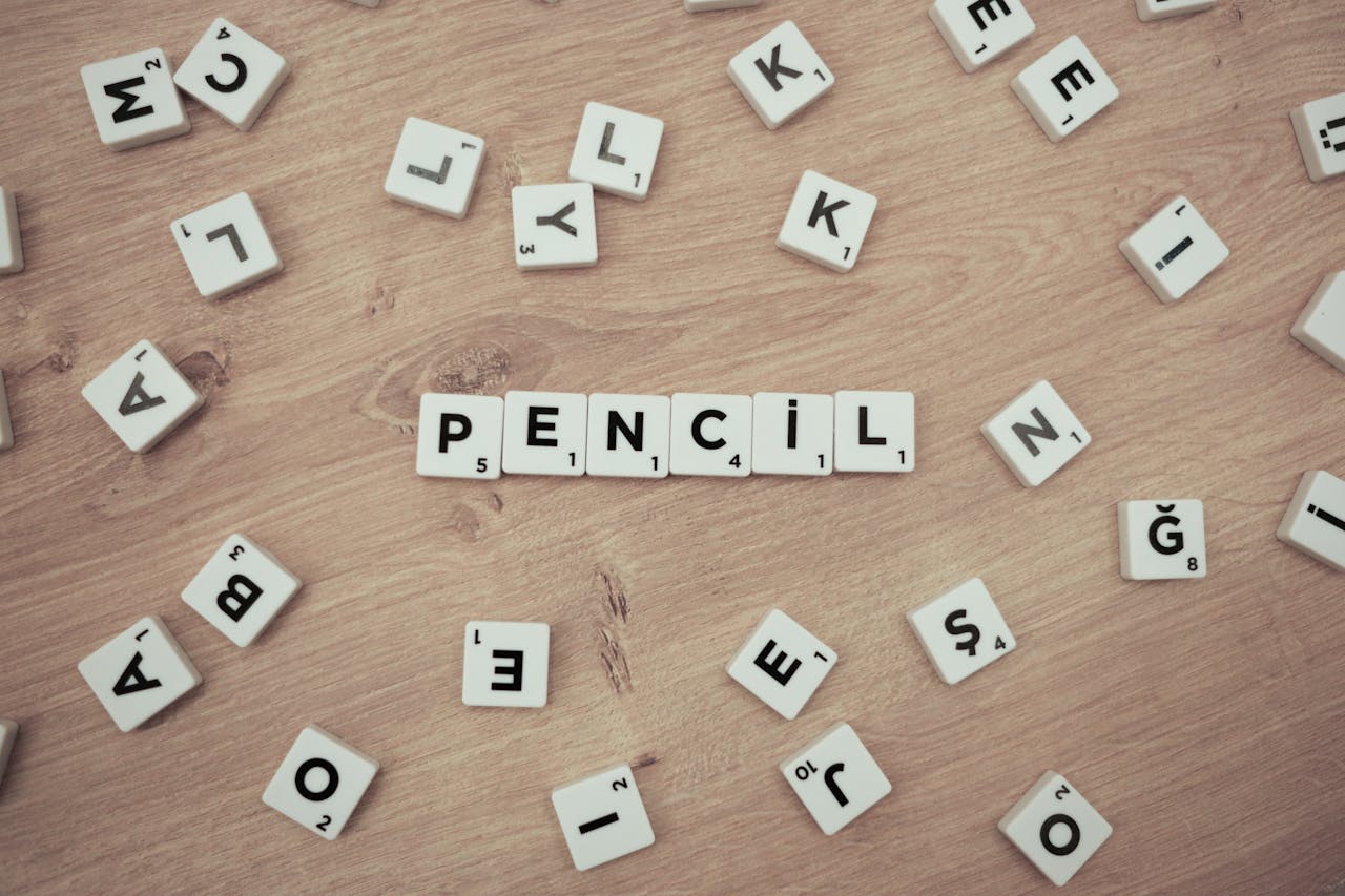 Wooden surface with scattered Scrabble tiles forming the word 'Pencil'.