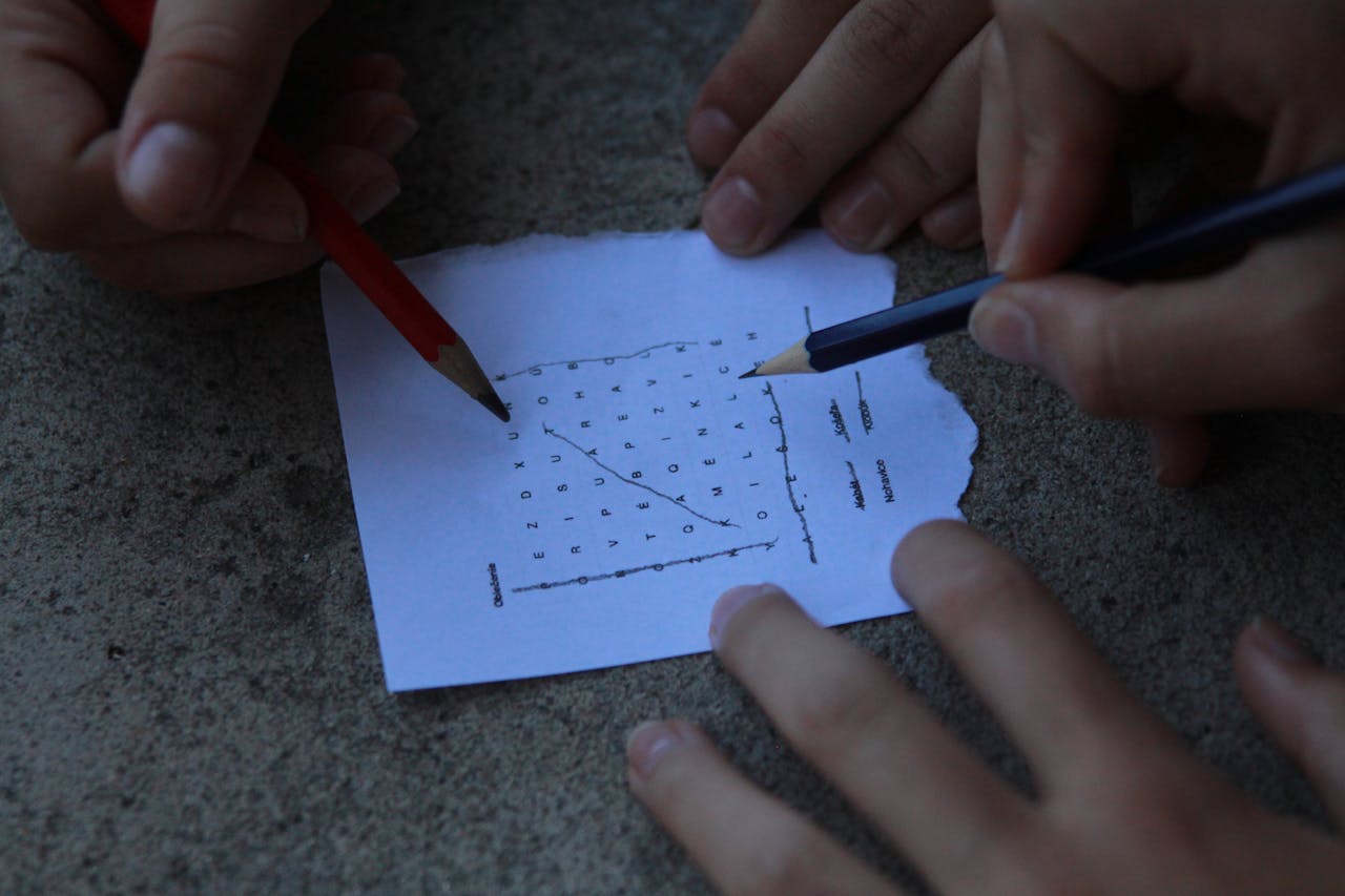 Close-up of hands using pencils to solve a word puzzle on paper.